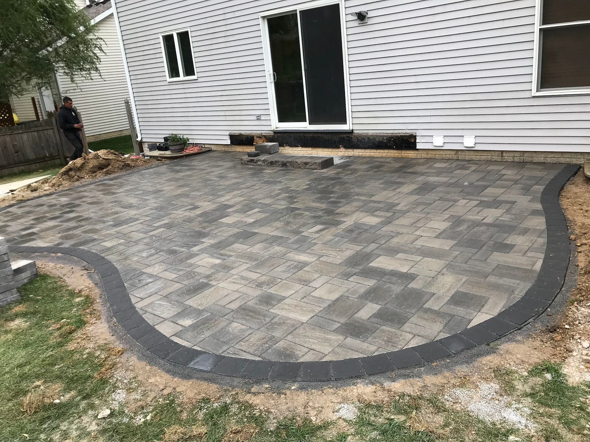 Patio with grill, table, and chairs. Paved with gray bricks, surrounded by mulch and a fence.