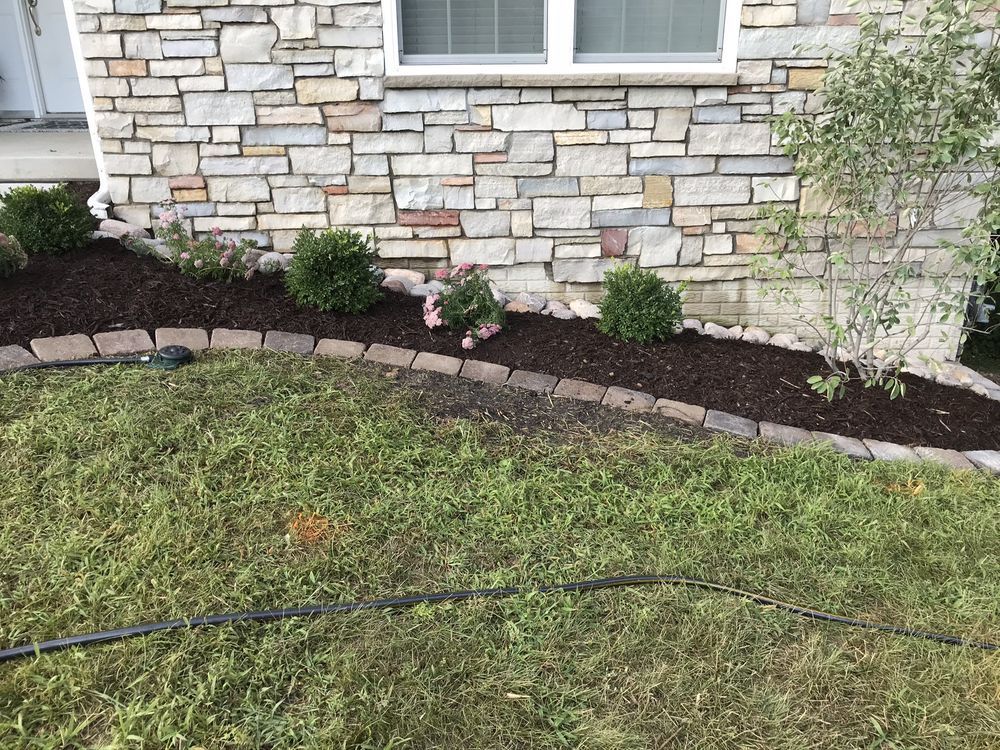 Landscaped flower bed with stone border, stone exterior wall, green grass, and bushes.