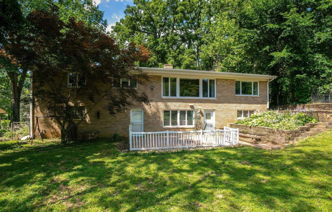 Brick two-story house with white trim, small deck, and lush greenery in a backyard setting.