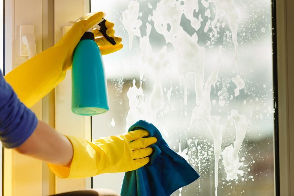 A person wearing yellow gloves is cleaning a window with a cloth and spray bottle — South Pacific Cleaning in Edmonton, QLD