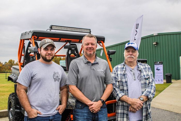 Three men stand in front of an off-road vehicle with an orange roll cage. Cloudy day, outside.