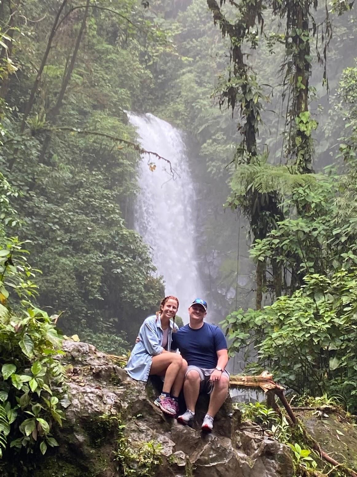 Two people sitting on a rock in front of a tall waterfall in a lush forest