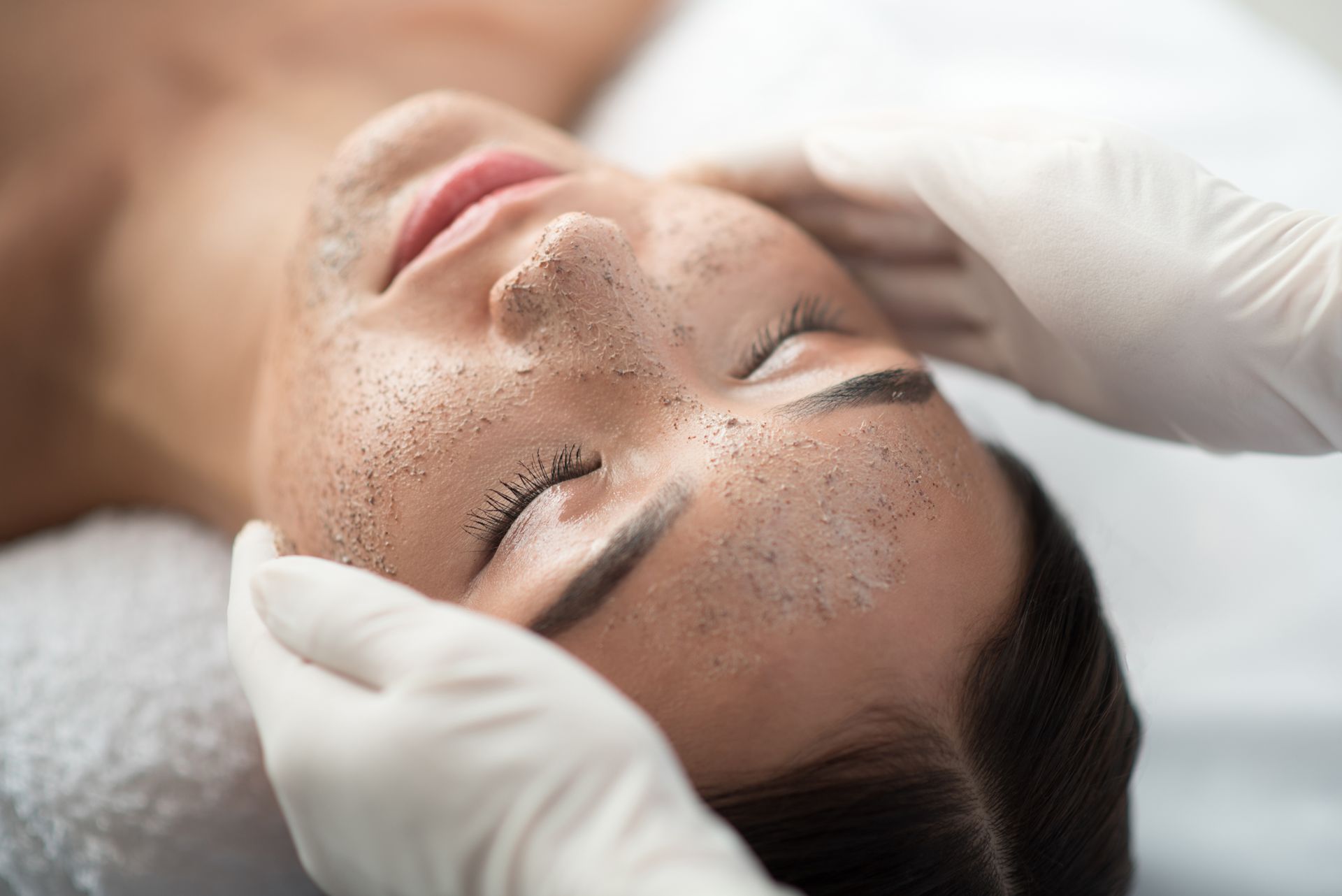 Person receiving facial scrub treatment, eyes closed, hands in white gloves.