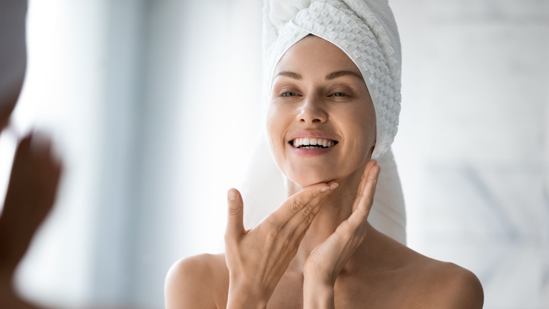 Woman smiles, looks in mirror, touches face with hands; towel on head.