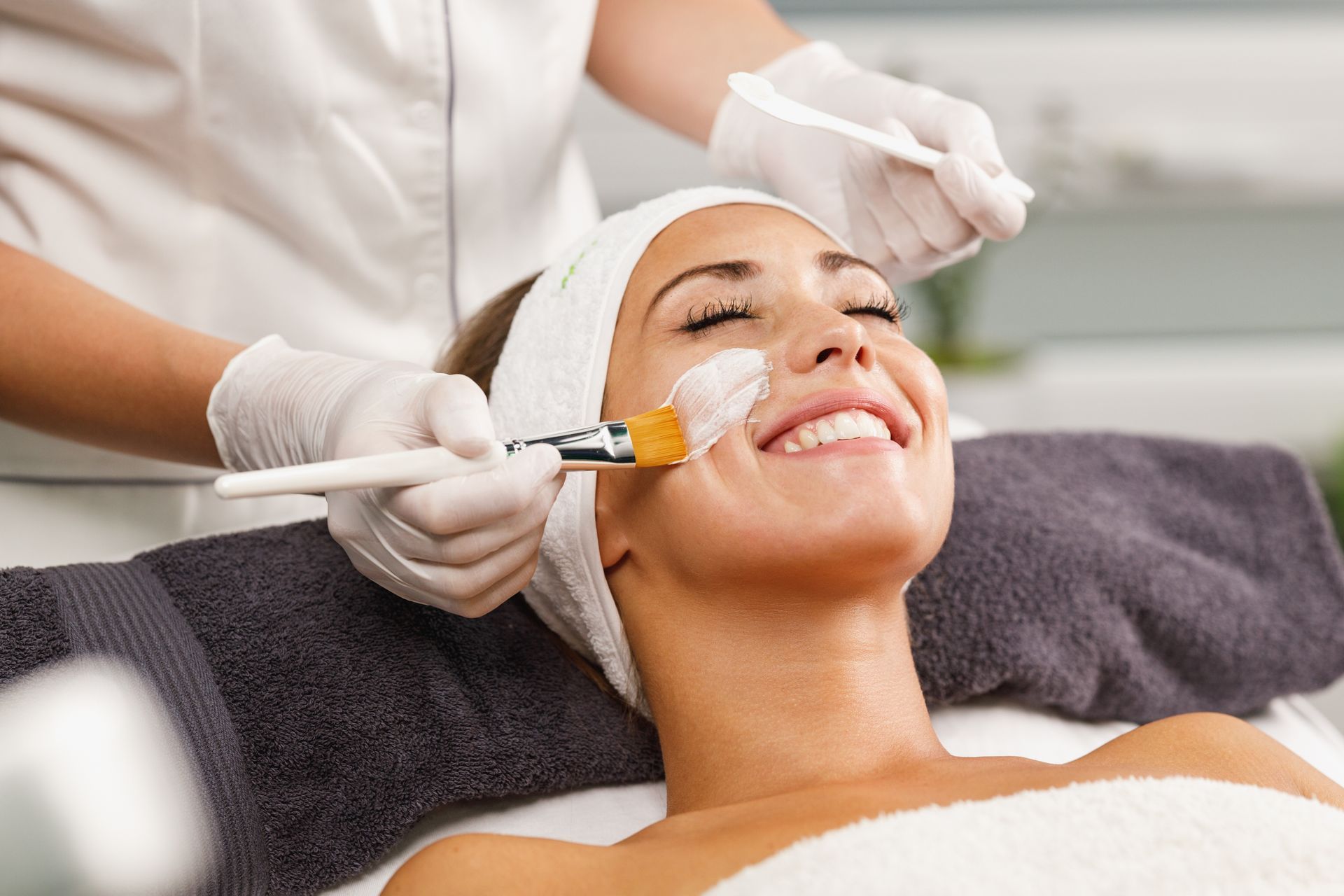Woman receiving facial treatment, smiling, with mask being applied by gloved hands.