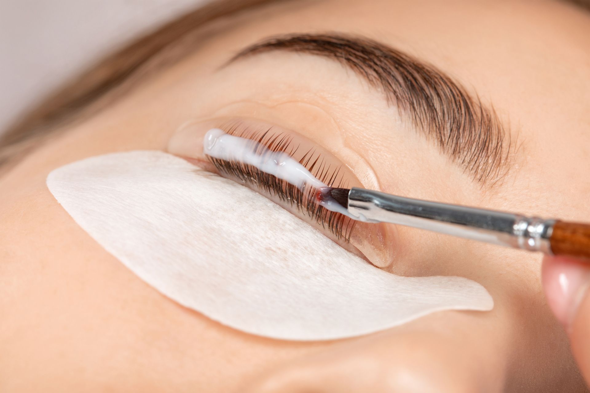 Person receiving a lash lift treatment. White cream applied to eyelashes with a brush, eye covered with a pad.