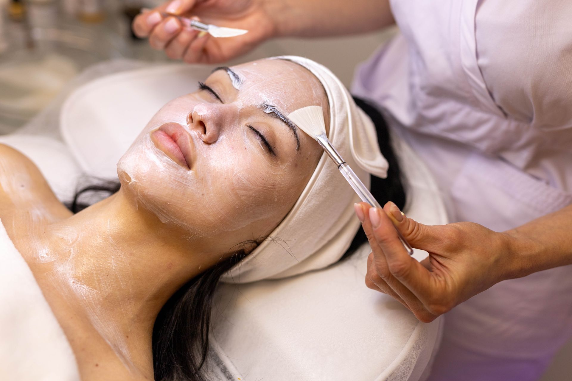 Woman receiving facial treatment, creamy mask applied to face by esthetician with tools.