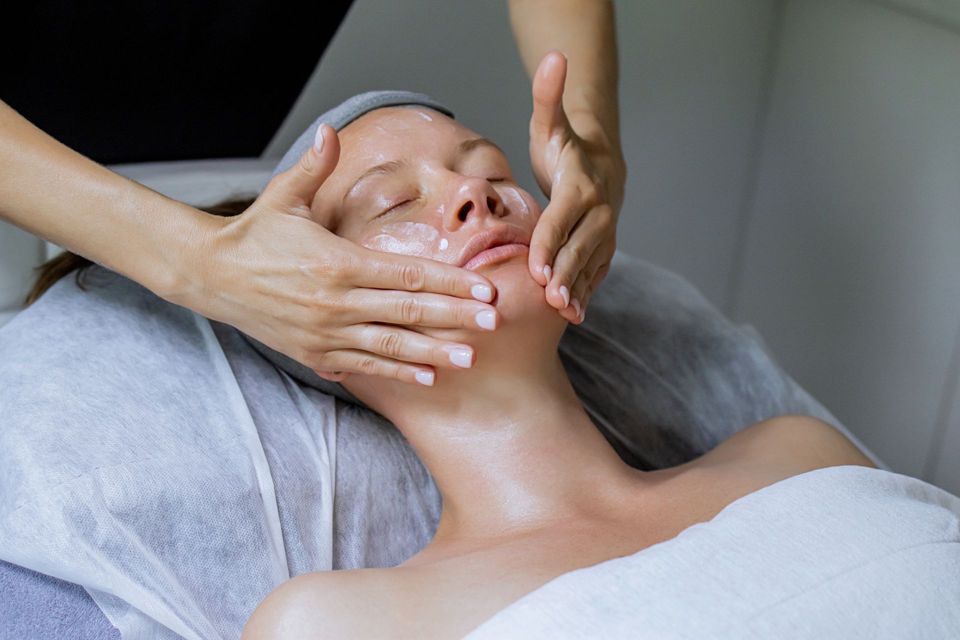 Person receiving facial massage; hands on face, cream visible; lying on white sheet.
