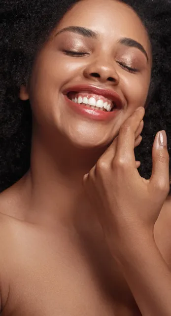 Smiling person with closed eyes, touching their cheek with a hand, against a light gray background.