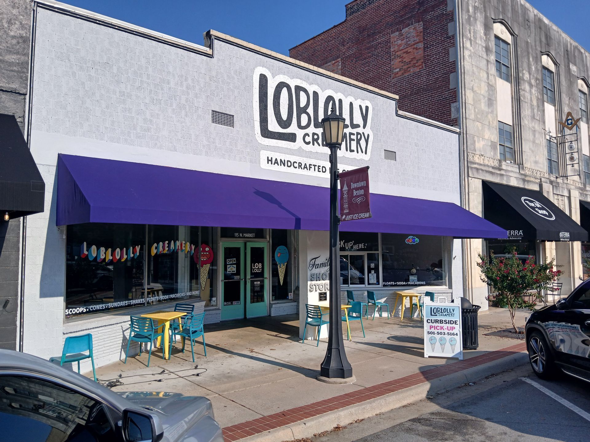 Purple-awning storefront with outdoor tables on a sunny street corner, seen from across the road