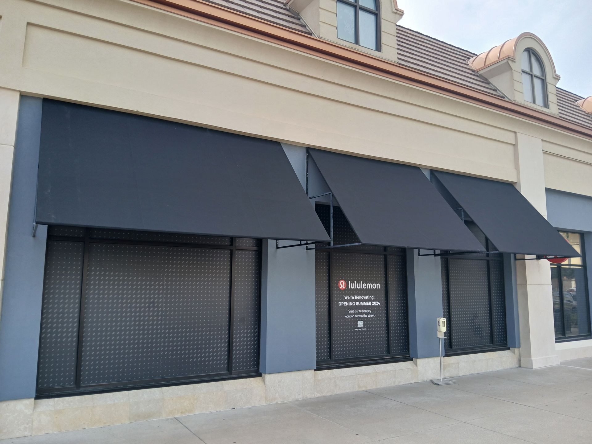 Closed storefront with black awnings and roll-down shutters on a beige building