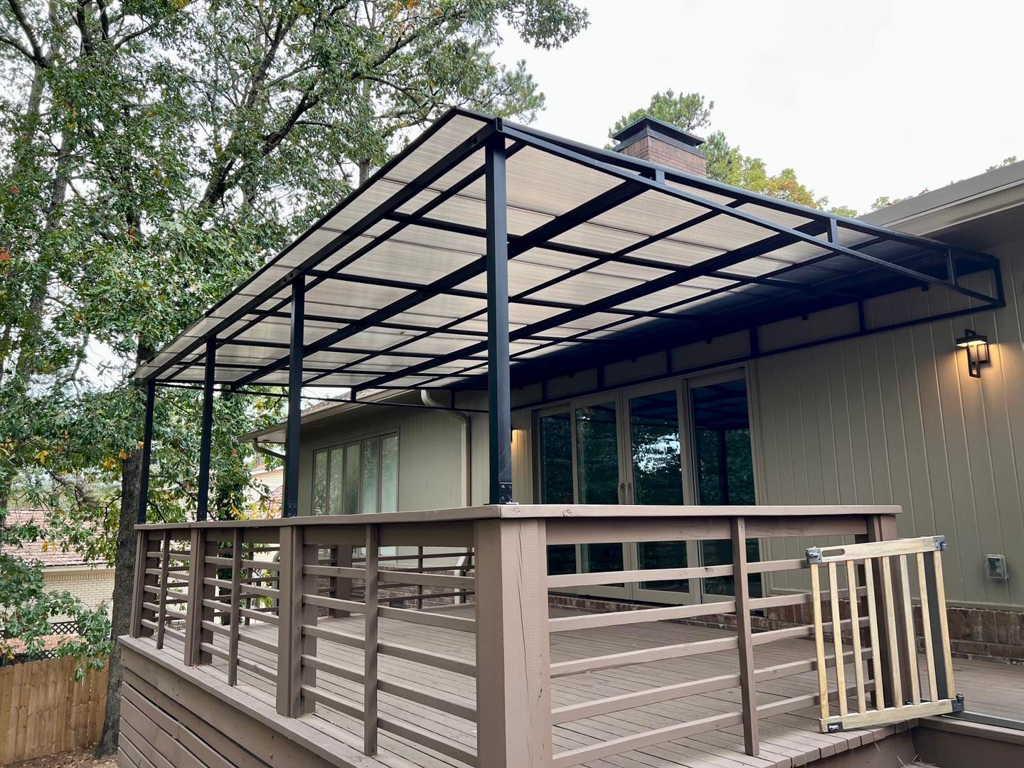 Wooden deck with pergola-style roof attached to a house, surrounded by trees and a railing.