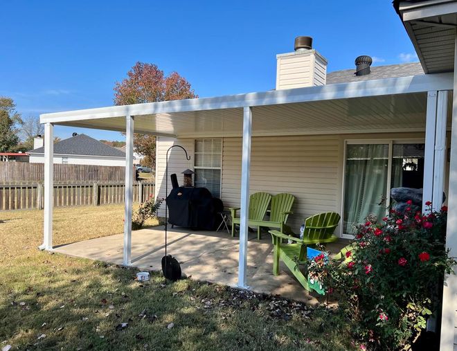 A white house with a covered patio with green chairs and a grill.