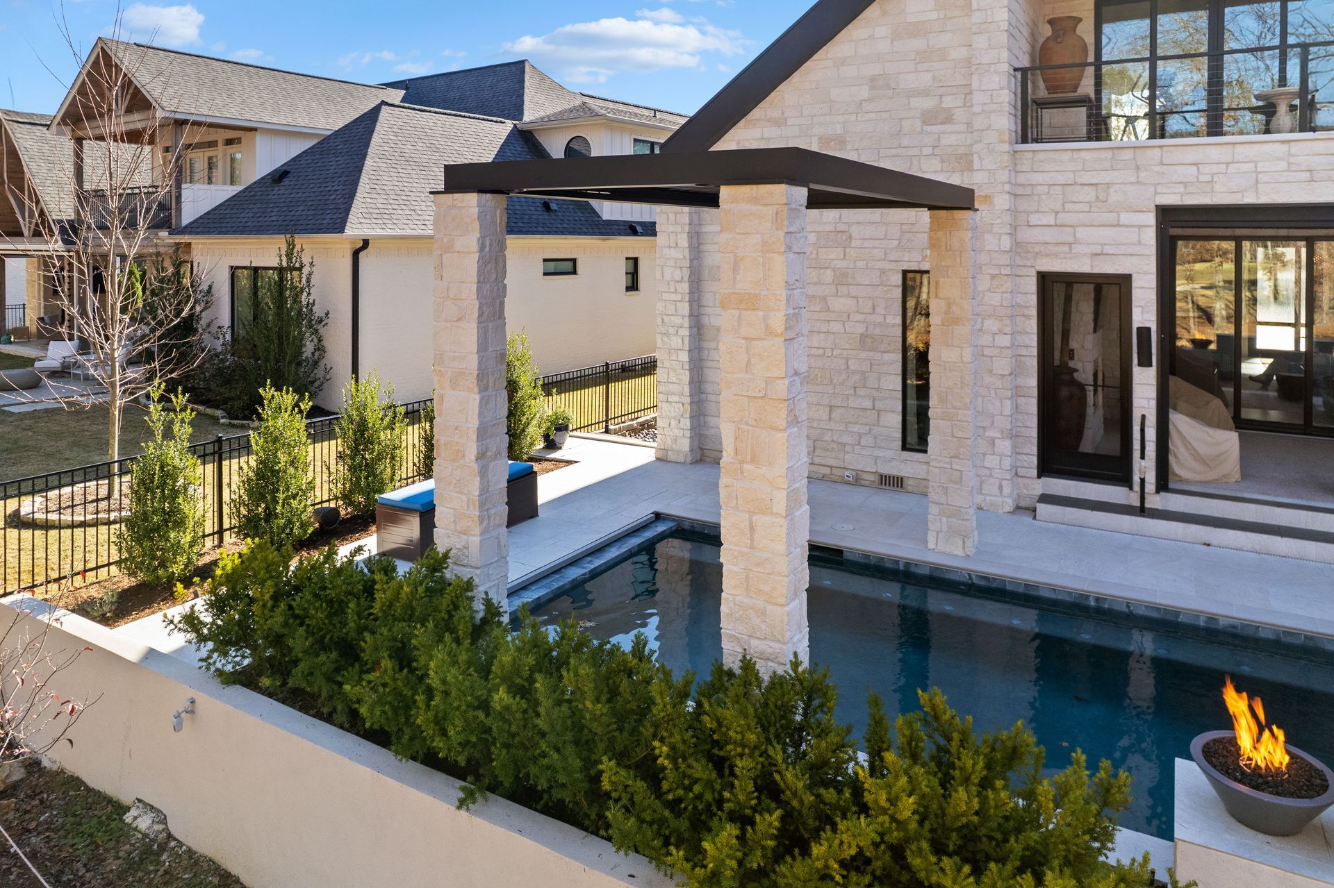Modern stone patio with pergola, pool, and fire bowl beside a house in a landscaped backyard