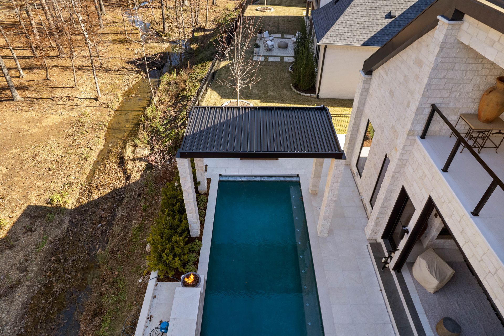 Aerial view of a narrow backyard pool beside a white house and dry hillside, with a small covered patio.