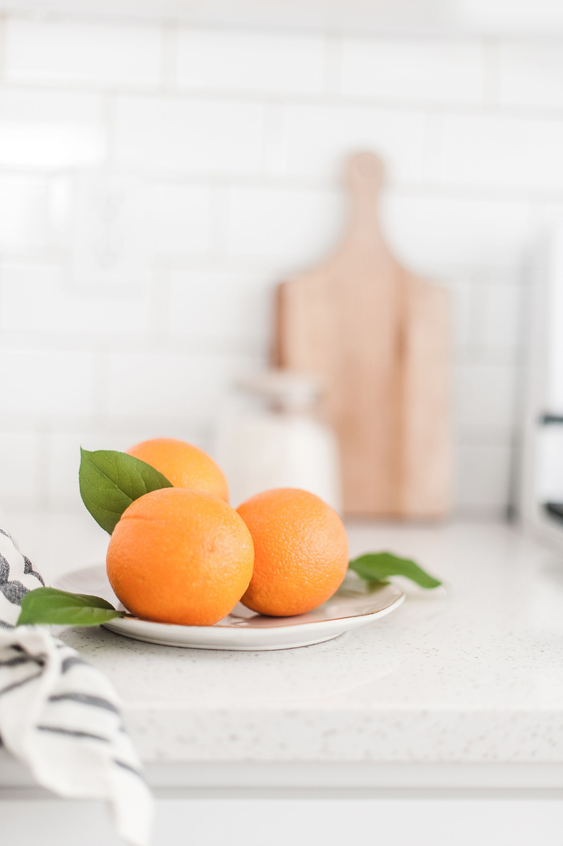 Oranges on a white plate on a kitchen counter, with a blurred cutting board in the background