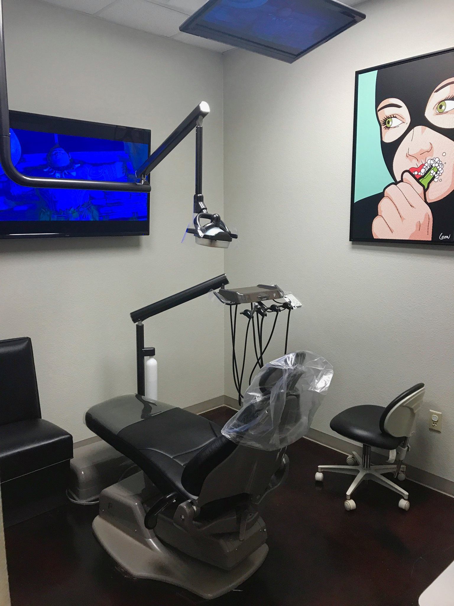 Modern dental exam room with chair, overhead light, wall screen, and colorful artwork.