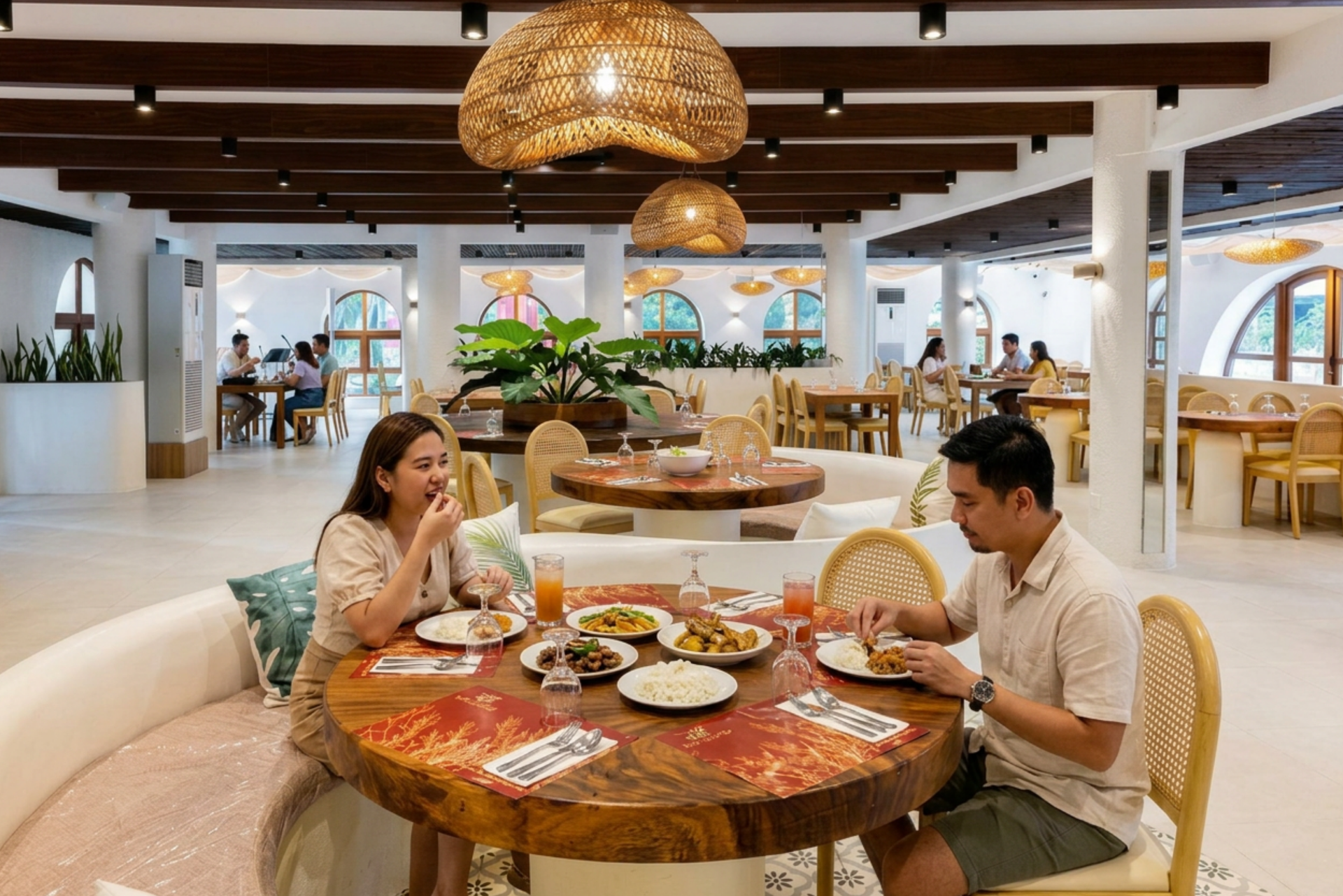 Two people dining at a round table in a bright, modern restaurant with woven pendant lights and ocean views