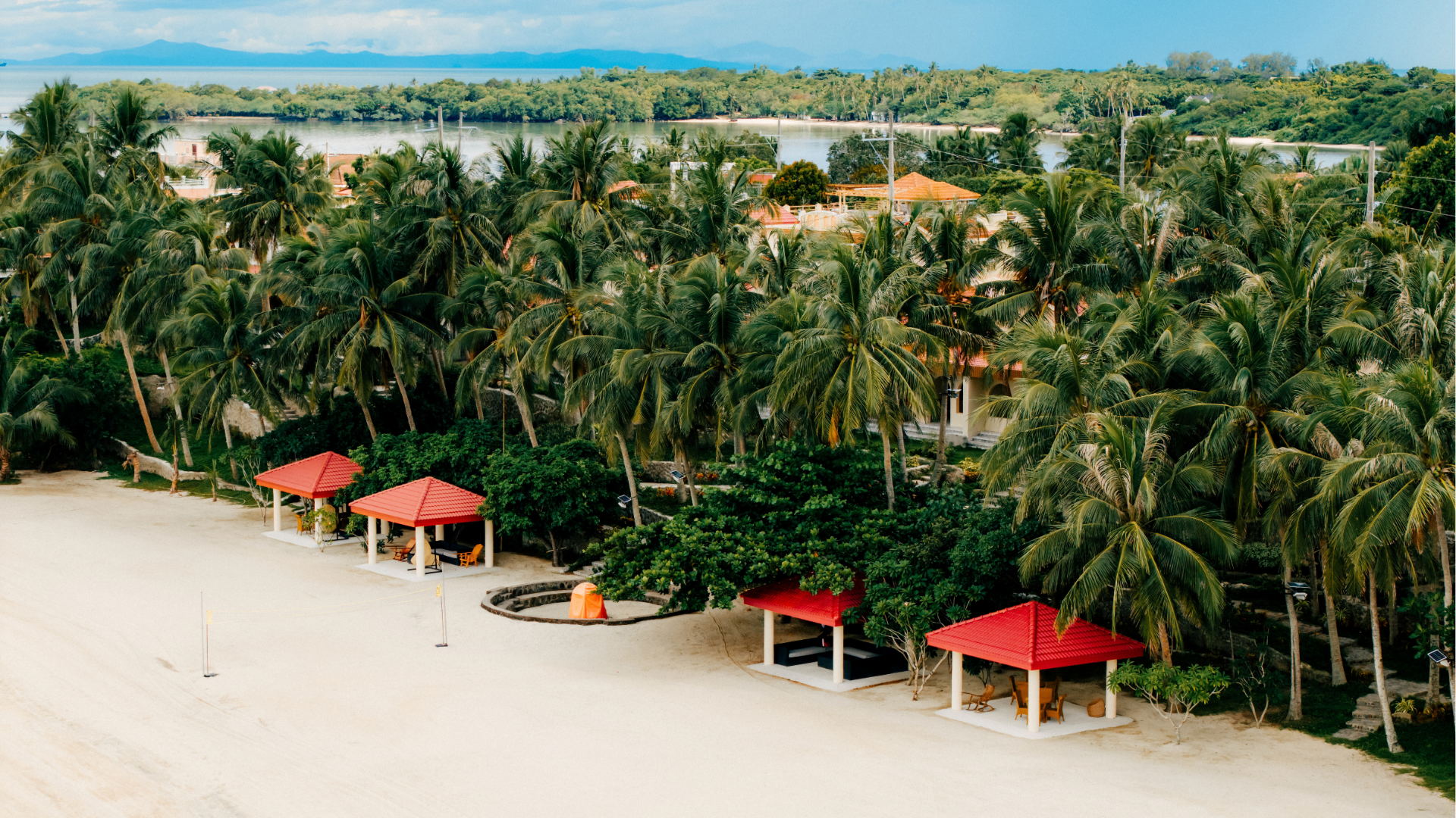 Sandy beach with red-roofed shelters and rows of palm trees beside calm blue water