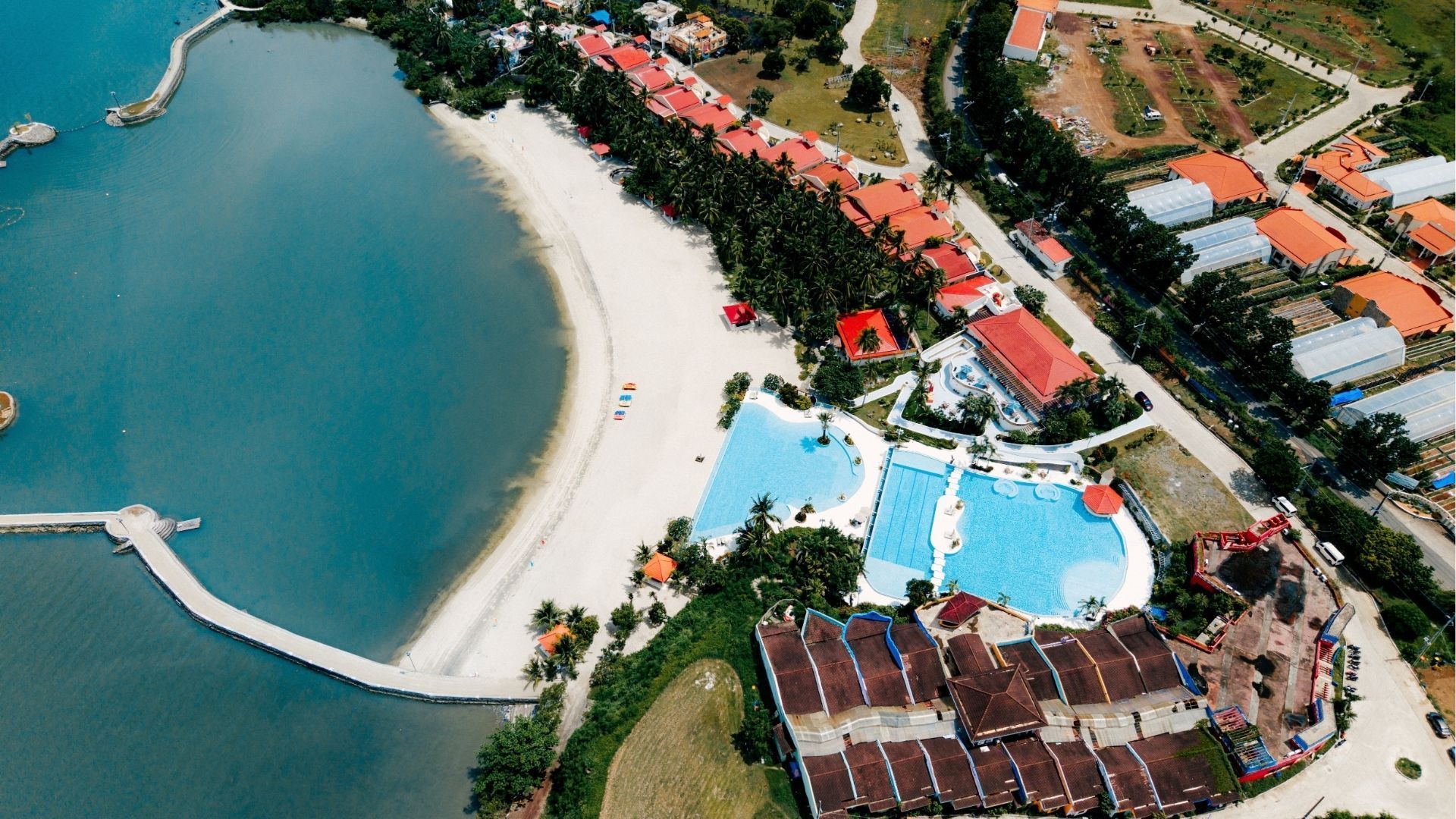 Aerial view of a beachfront resort with a curved sandy shore, blue pool, marina pier, and red-roofed buildings.