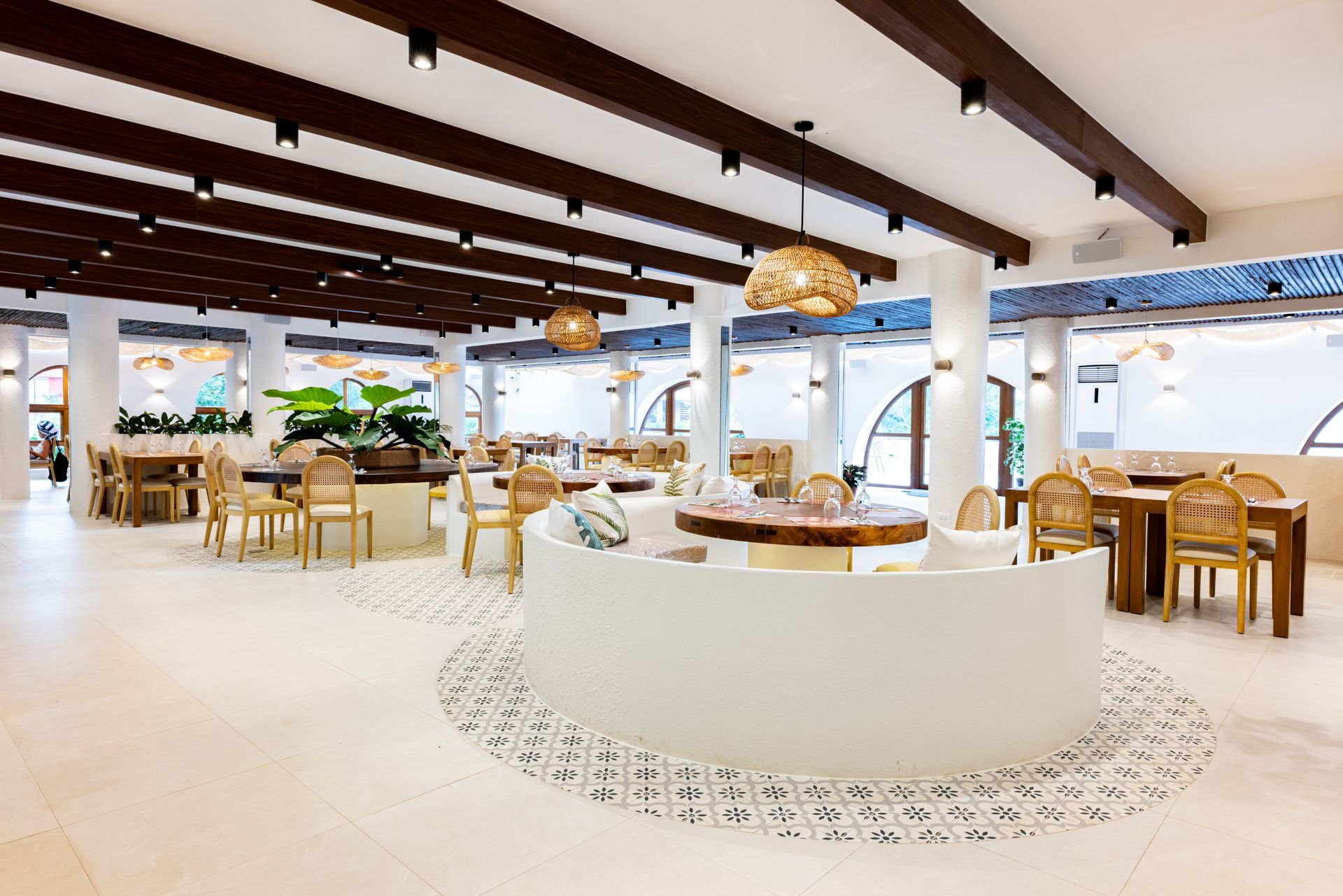 A modern, airy restaurant interior with neutral tones, rattan pendant lights, wooden beams, and curved seating.