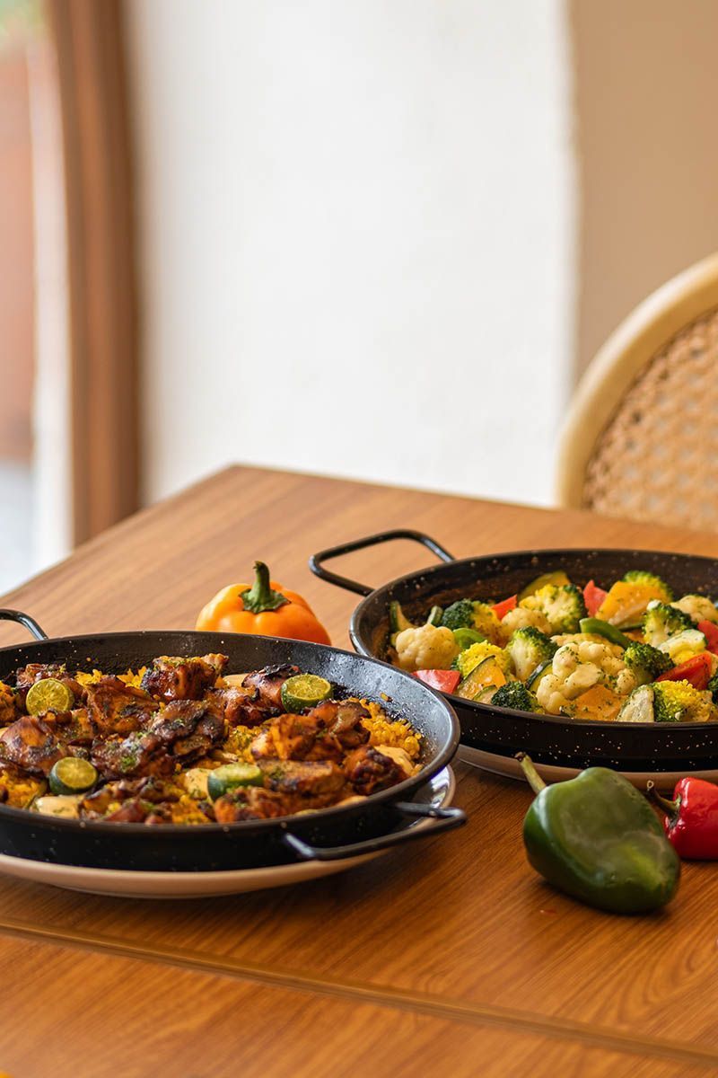 Two pans of roasted vegetables and food on a wooden table in a bright dining area