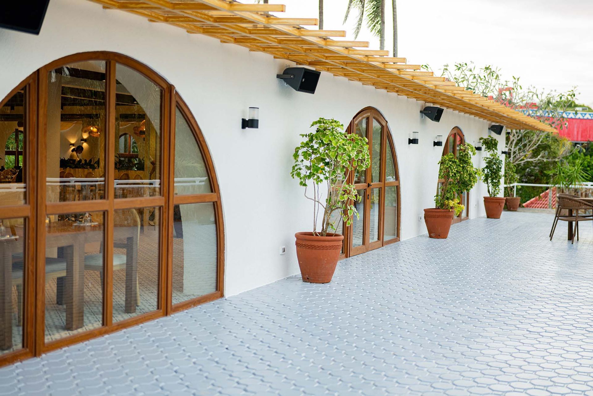 White courtyard with arched wooden windows, potted plants, and a patterned tile walkway under a sloped roof.