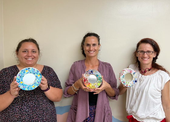 Three women smiling and holding colorful decorated plates against a plain wall.
