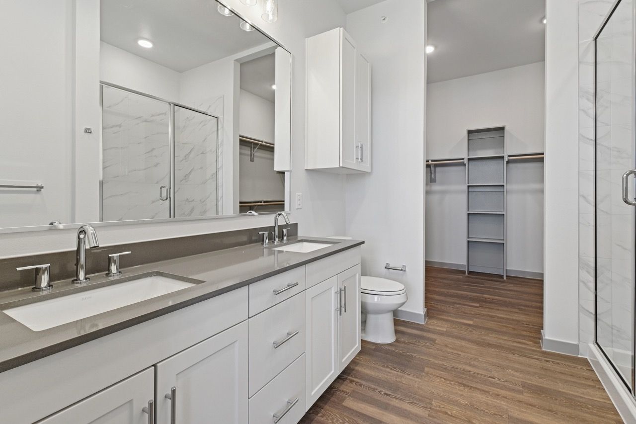 Modern bathroom with double-sink vanity, glass-enclosed shower, toilet, and open closet area.