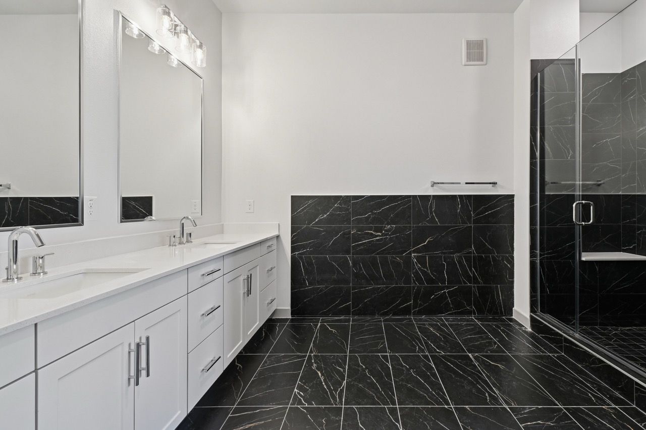 Bright bathroom with double vanity, white cabinets, large mirror, and glass shower with black marble tiles.