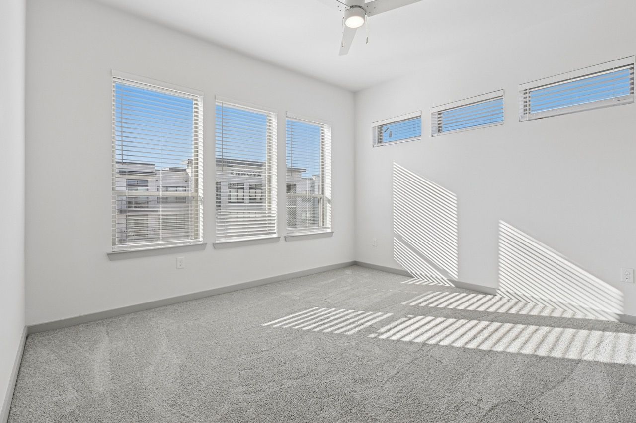 Bright, empty apartment room with white walls, carpeting, ceiling fan, and multiple windows with blinds.