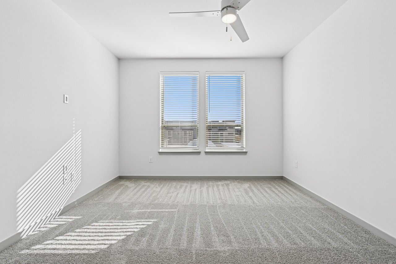 Empty white room with two windows, blinds, and a ceiling fan.