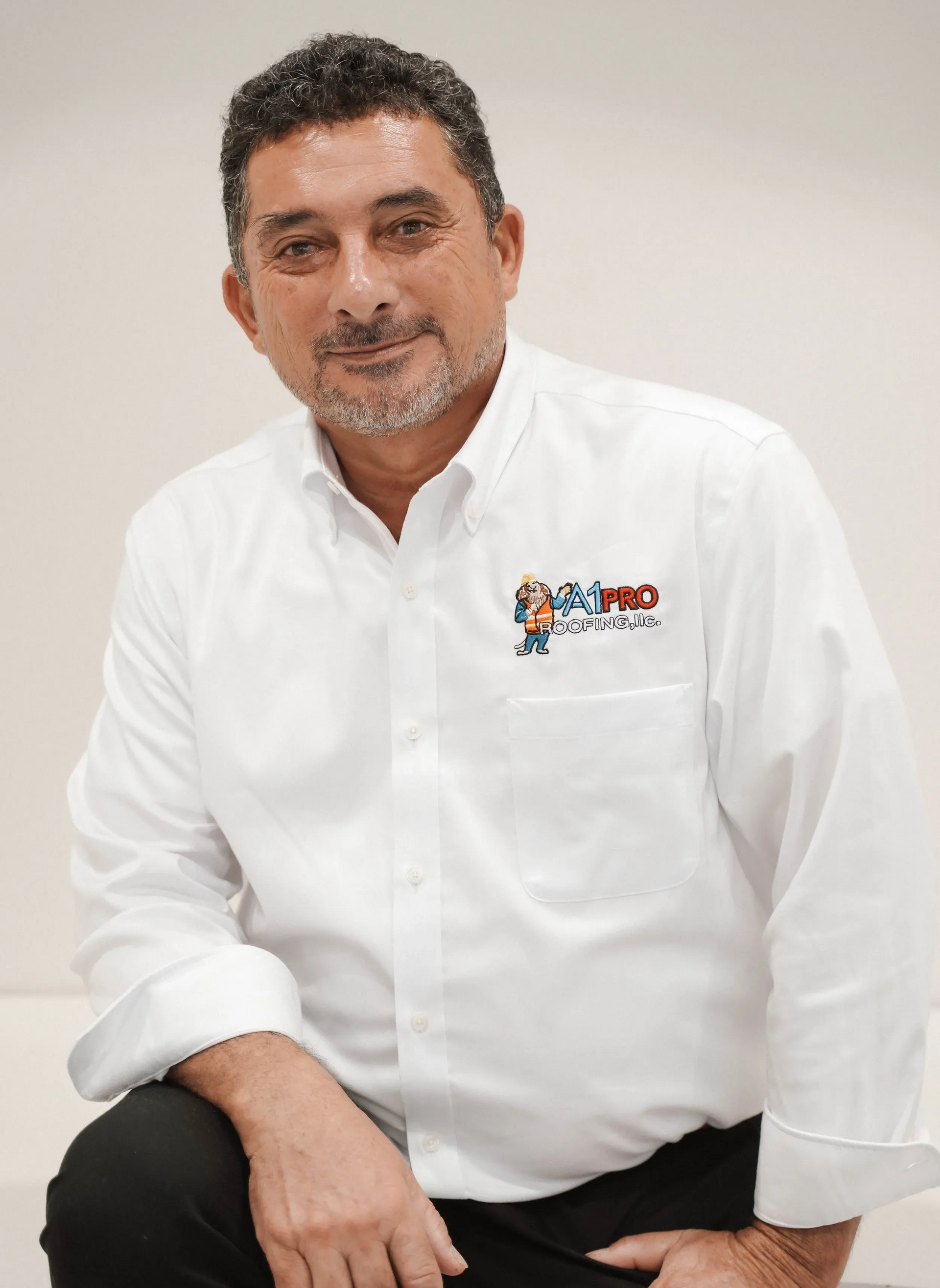 A man with a goatee wearing a white button-down shirt with a small colorful logo on his chest, looking toward the camera.