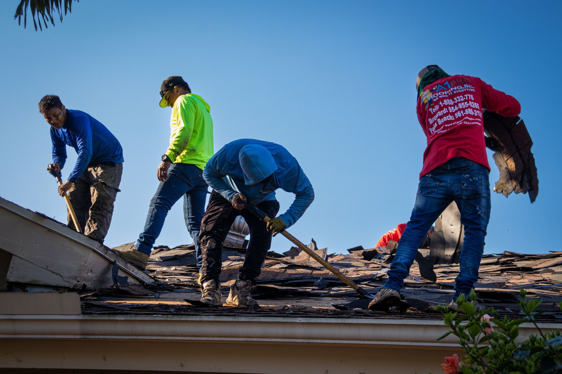 A person in a blue shirt applies sealant around a plumbing vent pipe on a flat, dark roof with many circular fasteners.