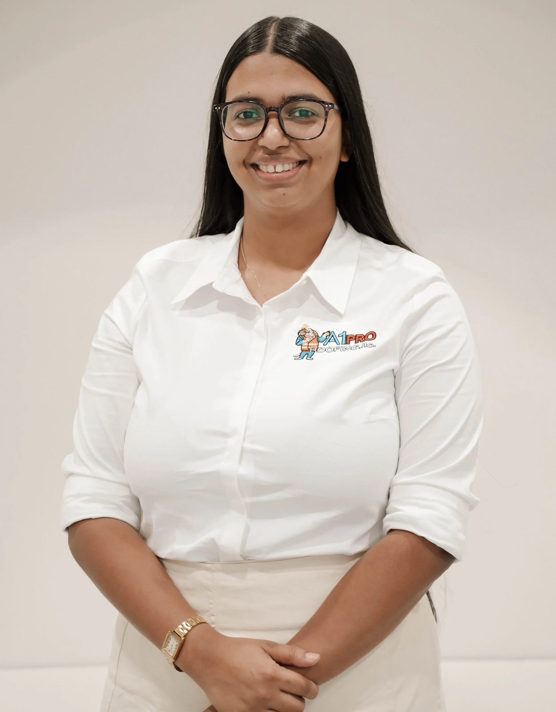 A smiling person with long dark hair and glasses wears a white collared shirt with a small logo, standing against a wall.