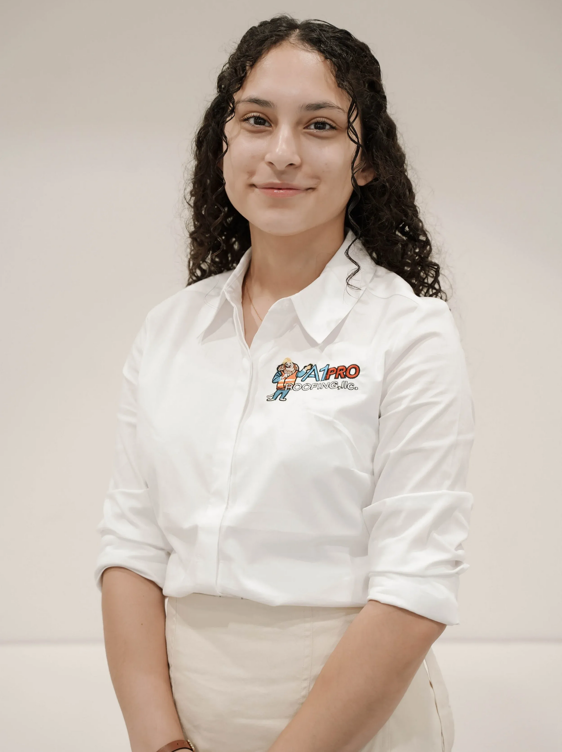 A person with curly dark hair wearing a white button-down shirt with a small colorful logo, posing against a plain wall.