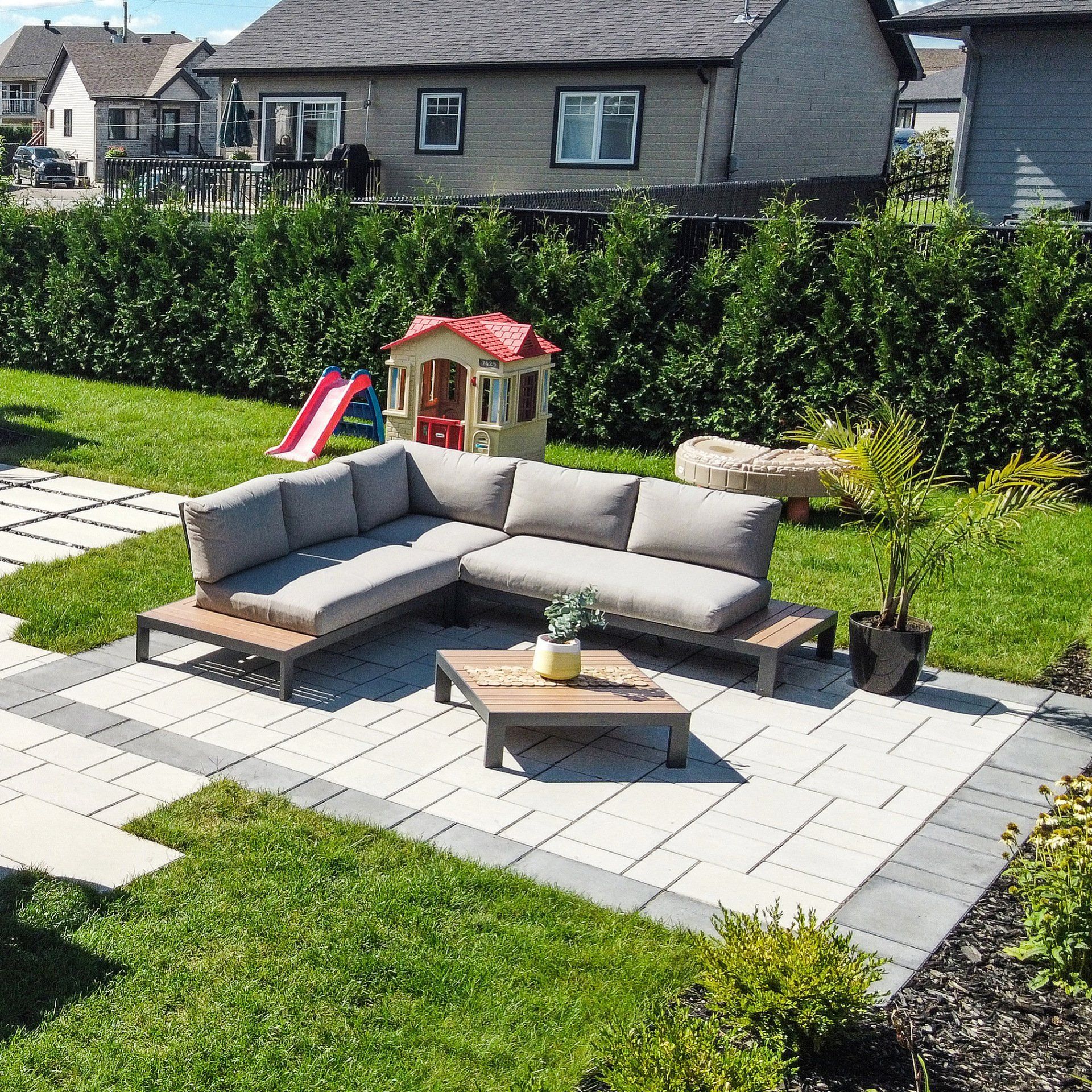 Canapé d'angle gris en forme de L avec table basse assortie sur une terrasse dans un jardin gazonné avec une cabane pour enfants.