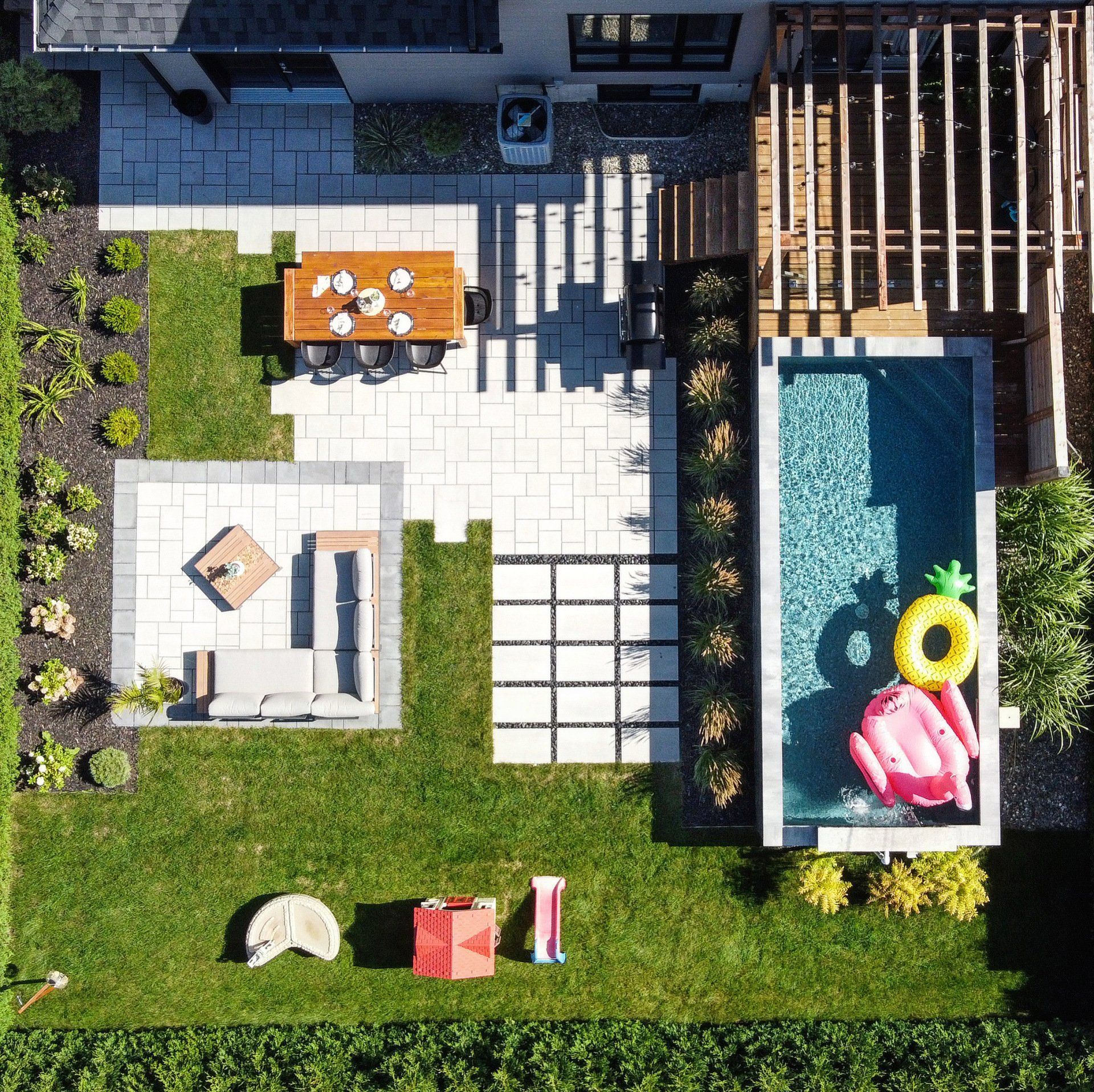 Vue aérienne d'une cour arrière résidentielle avec patio, coin repas, salon, piscine et pelouse avec jeux d'enfants.