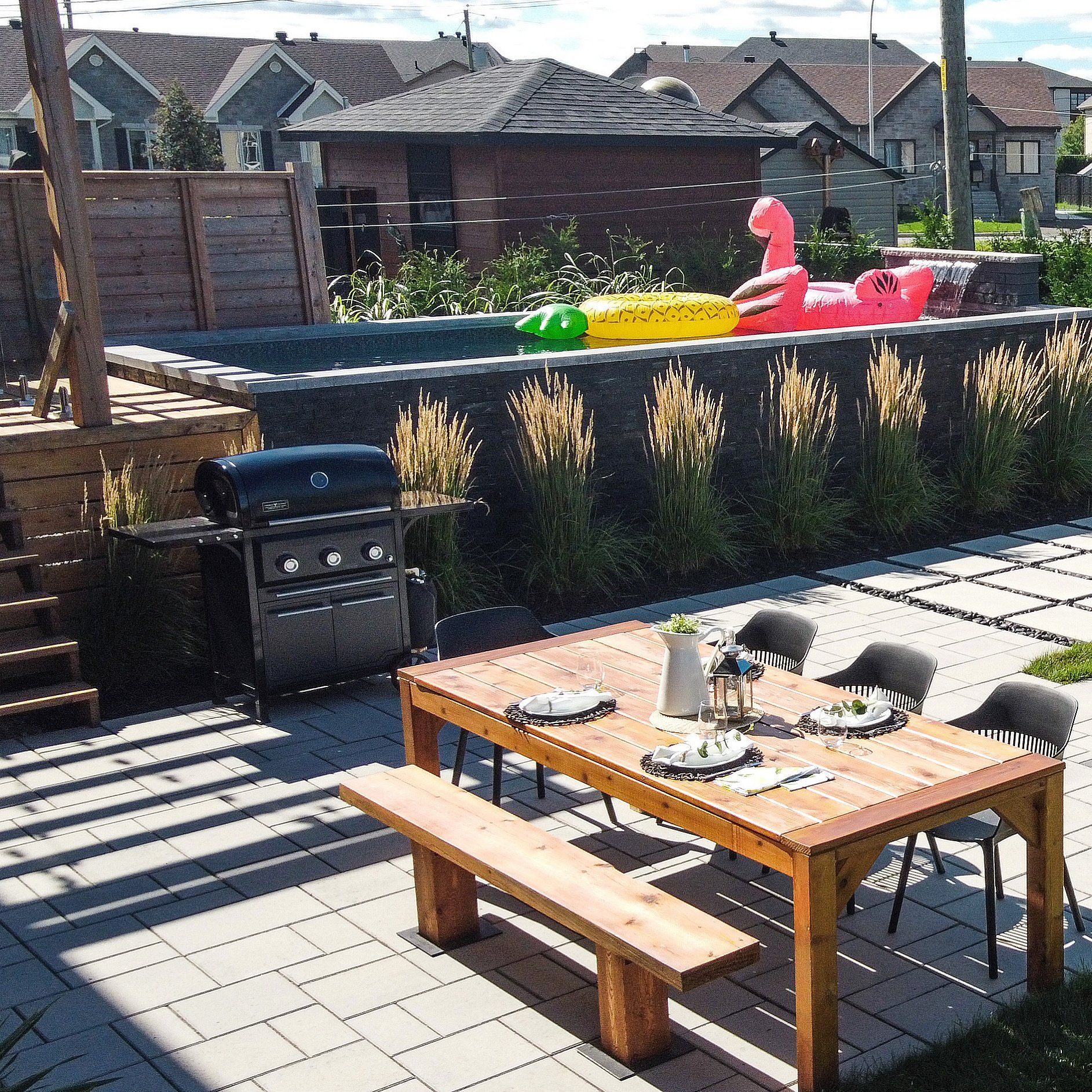 Une terrasse dans le jardin avec une table et un banc en bois, un barbecue à gaz noir et une piscine surélevée agrémentée de jeux gonflables colorés.