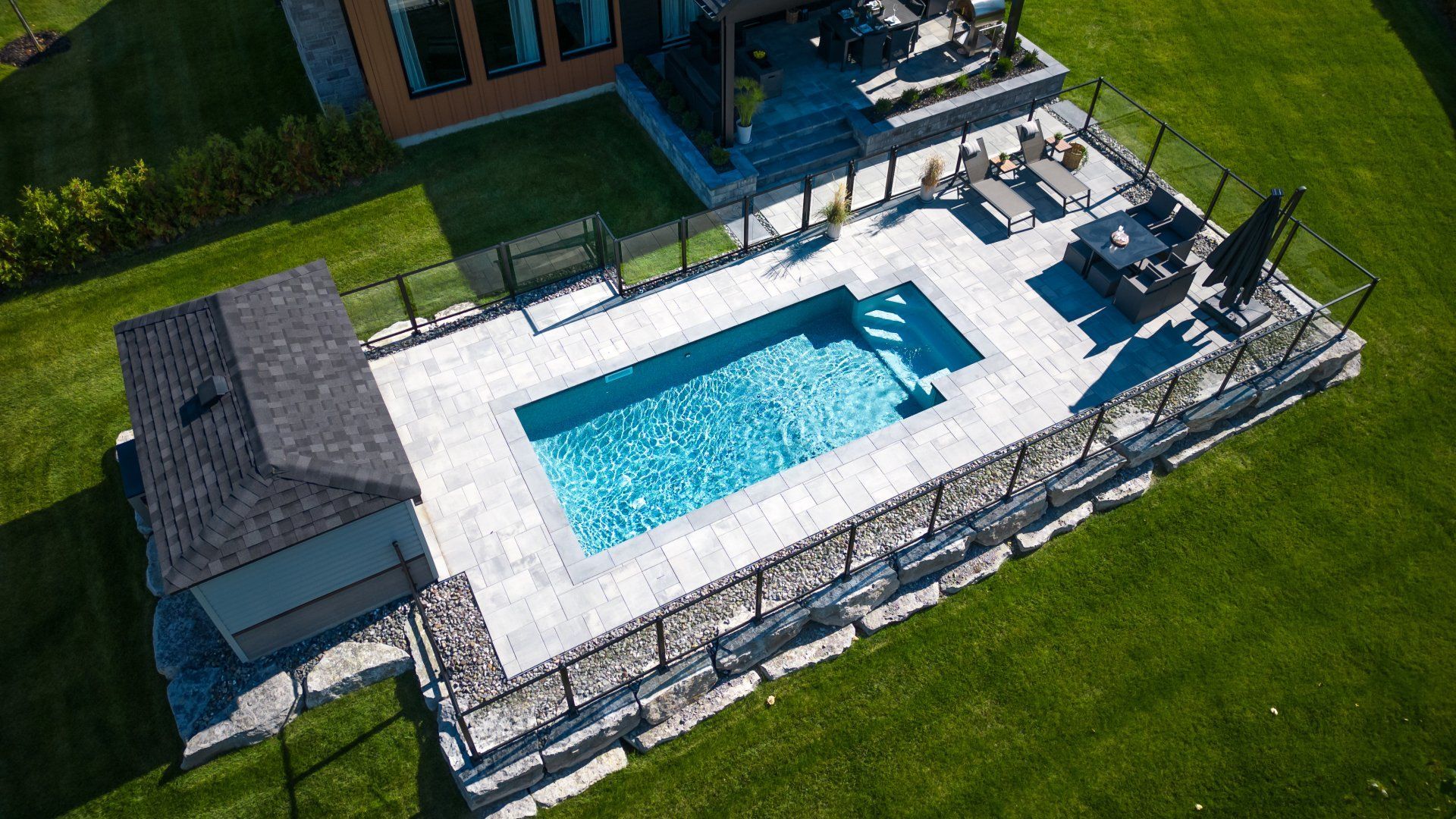 Vue aérienne d'une piscine rectangulaire dans un jardin, entourée d'une terrasse en pierre, d'une clôture en verre et d'un petit abri de jardin adjacent.