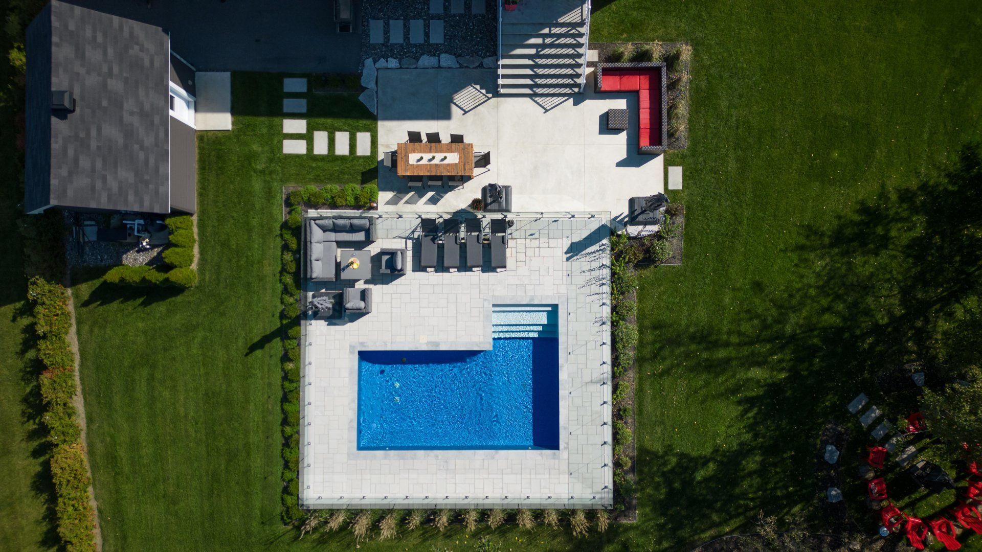 Vue aérienne d'un jardin avec une piscine rectangulaire, du mobilier de patio, un coin repas et un foyer sur une terrasse en pierre.