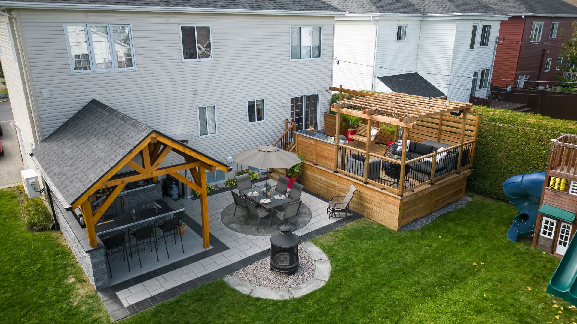 Terrasse arrière avec coin repas couvert, terrasse en bois surélevée avec pergola et foyer circulaire en pierre.