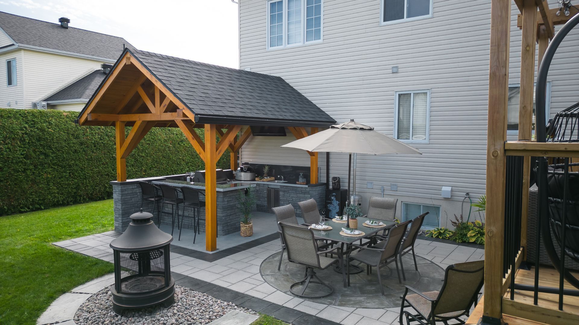 Terrasse arrière avec cuisine extérieure couverte, bar en pierre, table à manger avec parasol et foyer sur une pelouse.
