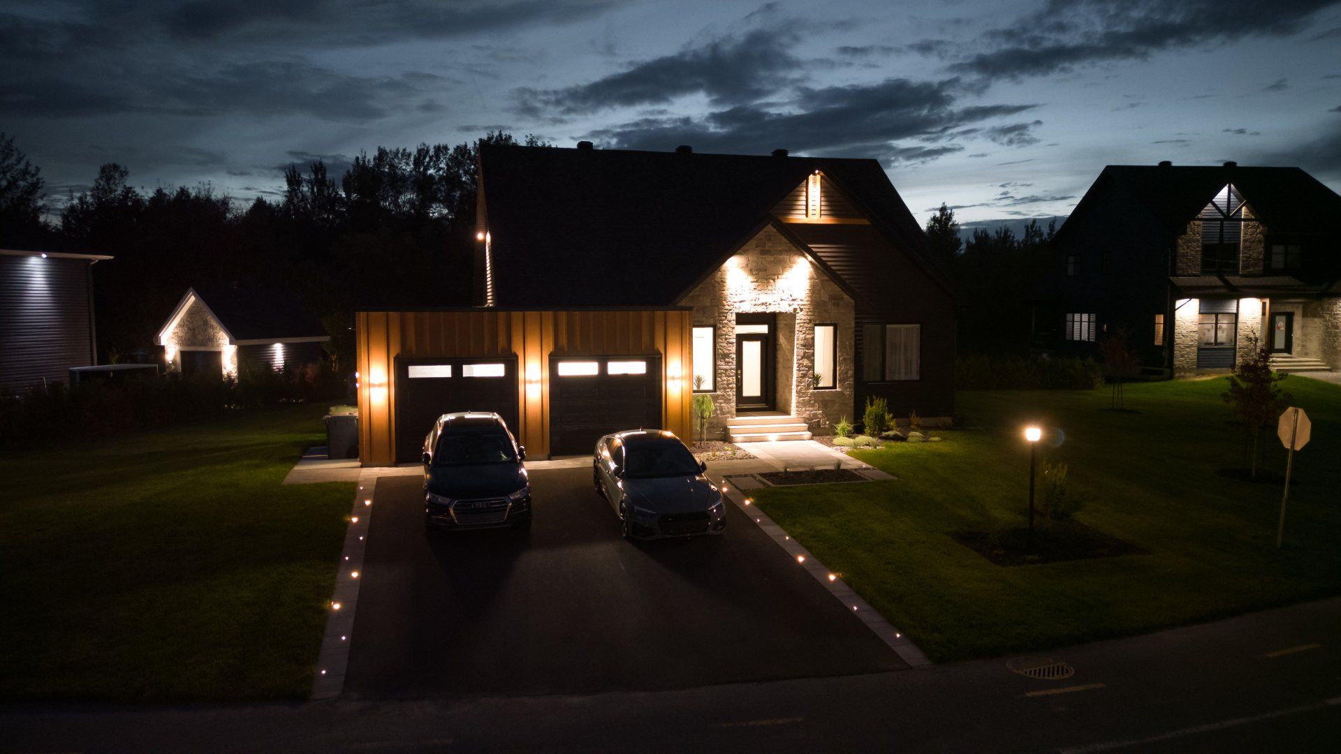 Une maison moderne à deux étages, la nuit, avec un éclairage extérieur d'accentuation, une allée pavée pouvant accueillir deux voitures et une pelouse.