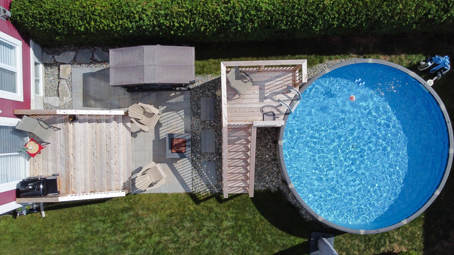Vue aérienne d'un jardin avec une piscine hors sol circulaire, une terrasse en bois, du mobilier de jardin et une pelouse environnante.