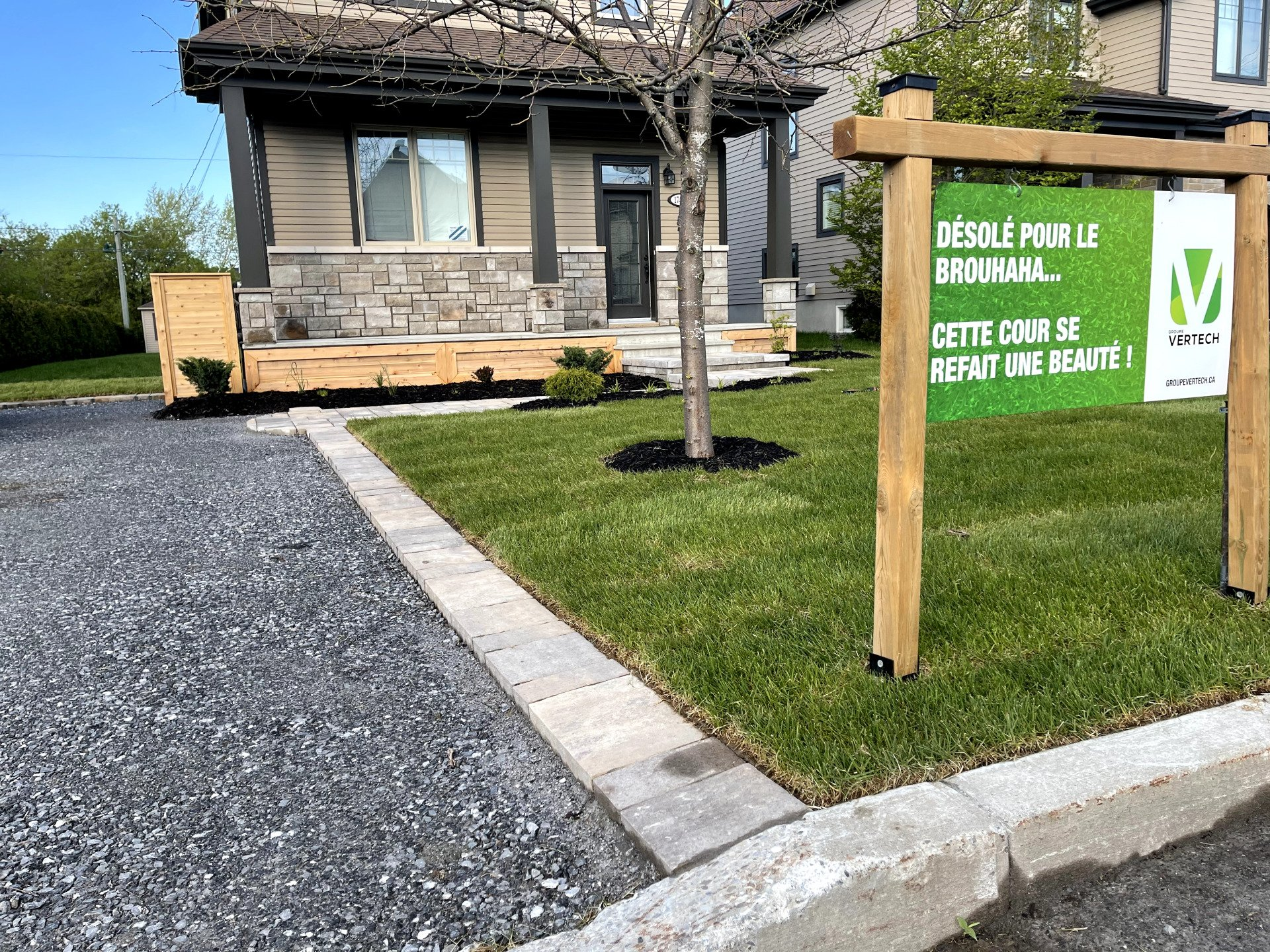 Une cour avant aménagée avec une allée en gravier, une bordure en pierre et un panneau publicitaire en français sur un cadre en bois.