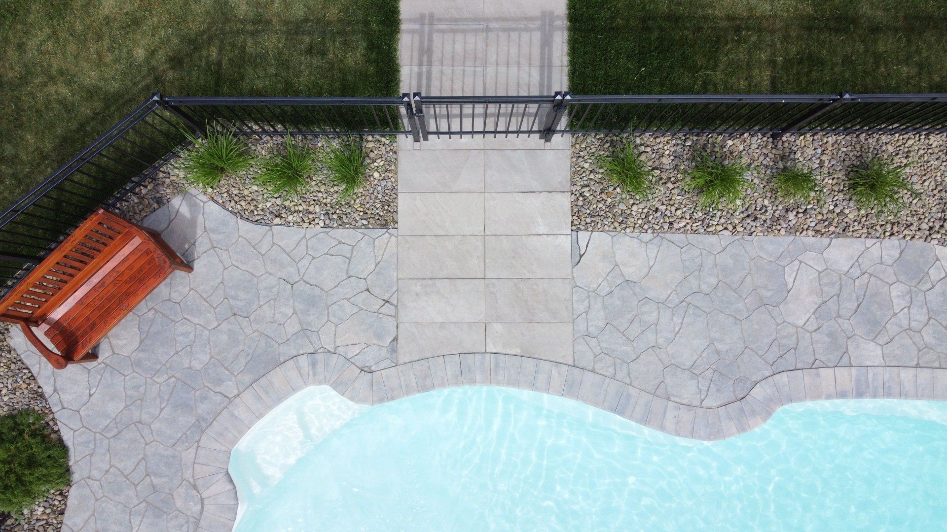 Une piscine à l'arrière d'une maison avec une terrasse en pierre, un banc en bois, un jardin clôturé et une allée en pierre menant à un portail.