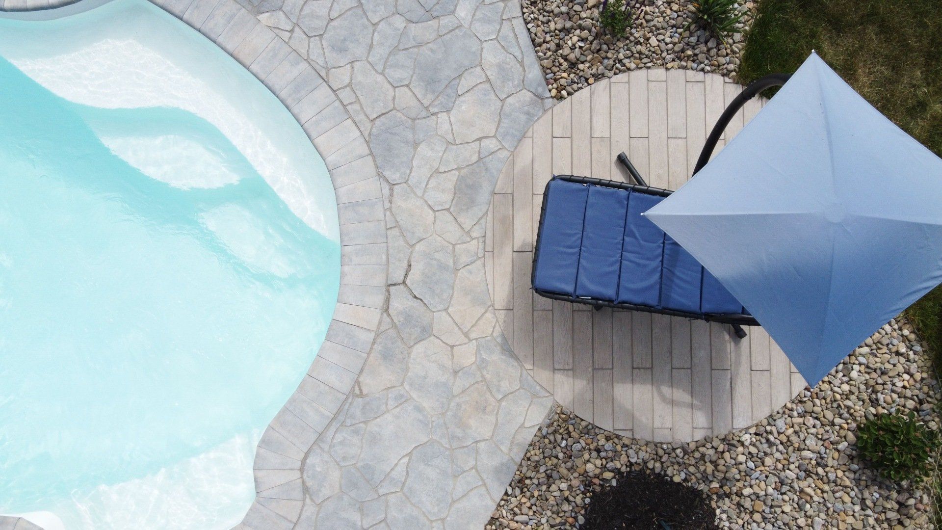 Un transat bleu sous un parasol bleu clair sur une terrasse circulaire jouxtant une piscine avec une terrasse en pierre.
