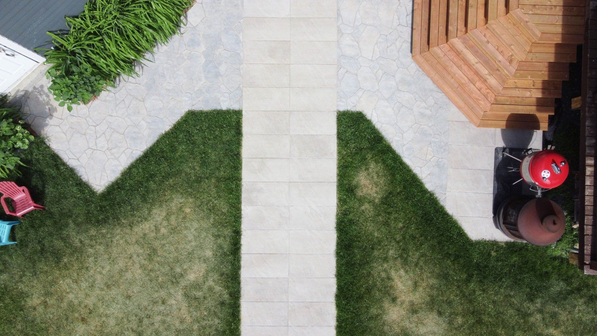 Vue aérienne d'un chemin de pierre bordé d'herbe, menant à une terrasse en bois avec un barbecue rouge et du mobilier de jardin.