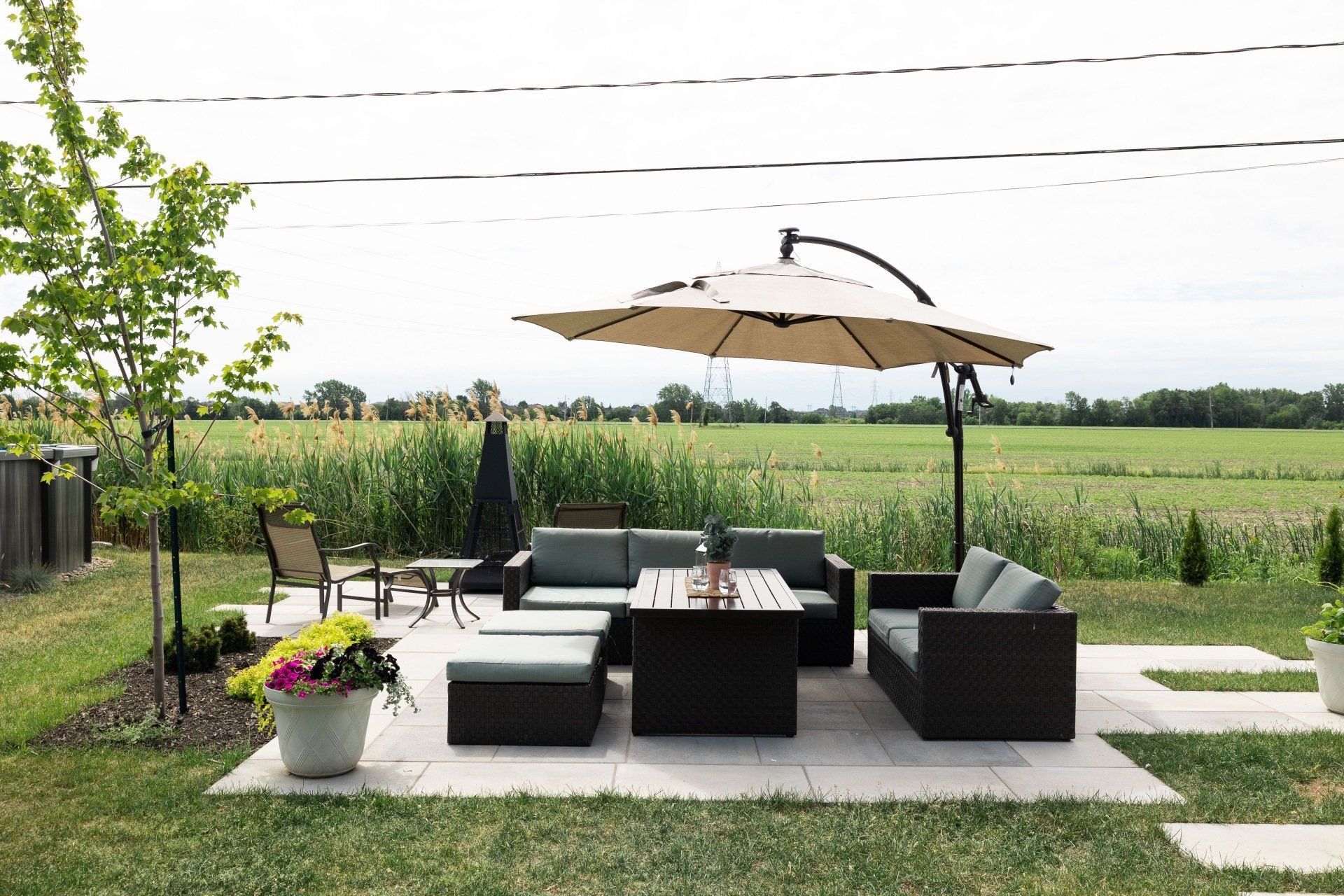 Un ensemble de patio comprenant un parasol beige, un canapé en osier foncé et une table basse assortie est installé sur des dalles de pierre donnant sur un champ.